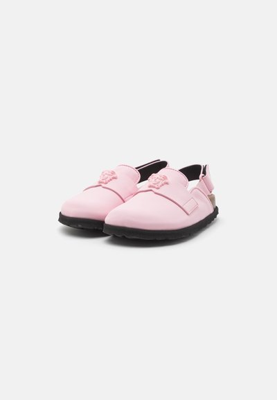 Rozā gumijas kurpes ar melnu zoli, ar plašu siksnu priekšpusē un lauvas logo detaļu augšpusē. Slingback dizains.
