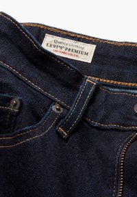 Mørkeblå jeans nærbilde som viser Levi's Premium-merket, gul søm, beltestropper og delvis synlig glidelås.