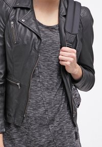 Giubbotto da moto in pelle nera con zip diagonale, accessori in metallo e una texture liscia, indossato sopra una maglia grigia melange. Cinghia nera visibile.