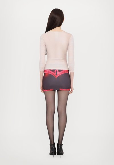 Durchsichtige Langarmbluse über einem dunklen Minirock mit rosa Spitzenverzierungen, kombiniert mit schwarzen durchsichtigen Strumpfhosen und hochhackigen Schuhen.