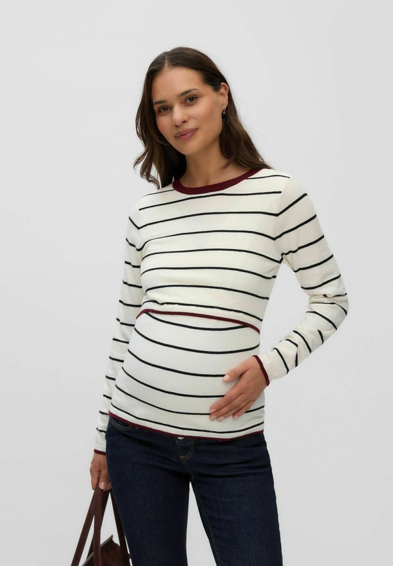 Femme enceinte portant un pull blanc à rayures et un jean foncé, tenant doucement son ventre et portant un sac marron devant un fond uni.