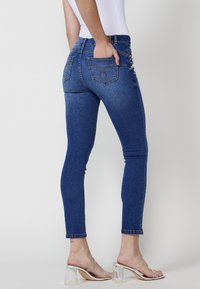 Skinny denim jeans in dark blue, featuring five pockets, reinforced seams, and decorative button accents along the left side. Textured fabric.