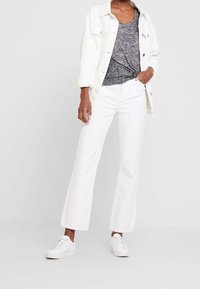 Persona che indossa pantaloni flair bianchi a vita alta, giacca di jeans bianca, t-shirt grigia annodata e sneakers bianche su uno sfondo chiaro e semplice.