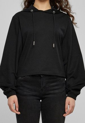 Personne portant un sweat à capuche court noir avec des cordons et des manches longues bouffantes, associé à un jean noir ajusté.