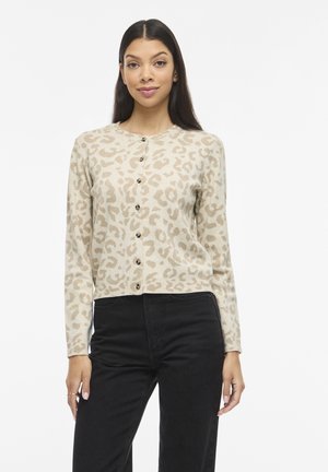 Mujer con un cárdigan beige con estampado de leopardo abotonado y pantalones negros de cintura alta, posando frente a un fondo blanco, sonriendo ligeramente.