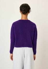 Vijolična, puhasta pletena jopica s spuščenimi rameni in skrajšanim robom, v kombinaciji s sivimi trenirkami, ki prikazuje sproščen in udoben videz.