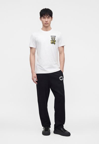 Νέος άνδρας φοράει λευκό t-shirt Moschino με γραφικό αρκουδάκι και μαύρη φόρμα Moschino, στέκεται μπροστά σε απλό φόντο.