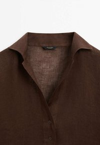 Chemise en lin marron foncé avec col, deux boutons visibles et une étiquette à l'intérieur de l'encolure sur fond blanc.