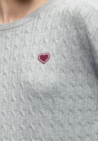 Pull gris clair en maille torsadée avec un petit écusson brodé en forme de cœur rouge sur la partie supérieure de la poitrine près de l'encolure.