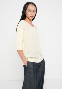 Bluză galben deschis cu mâneci scurte, decolteu rotund, textură moale, croială ușor lejeră și tiv drept, asortată cu pantaloni din denim închis la culoare.