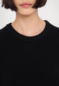 Femme avec une coupe courte noire type carré, portant un pull en maille côtelée noire, ne montrant que le bas du visage et le cou sur un fond uni.