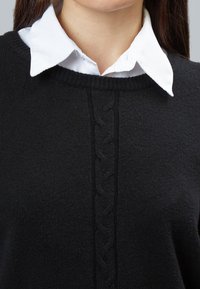 Schwarzer Pullover mit Rundhalsausschnitt, der ein strukturiertes Zopfmuster in der Mitte aufweist. Ein weißes, geknöpftes Hemd ist darunter sichtbar.