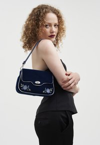 Mörkblå sammetsväska med blommig broderi, silverhårdvara, lockstängning och en enda rem. Rektangulär form med en strukturerad yta.