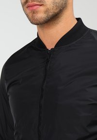 Svart bomberjacka med en slät, glänsande textur, ribbad krage och hel dragkedja fram. Har rena linjer och en minimalistisk design.