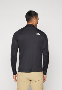 Zwarte lange mouwen shirt van lichtgewicht stof. Heeft een hoge kraag en het North Face-logo in het wit op de bovenrug. Textuur.
