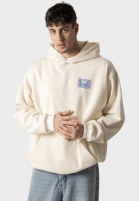 Felpa con cappuccio color crema, con un patch grafico che presenta un design di montagna e del testo, realizzata in morbido tessuto e dalla vestibilità rilassata.