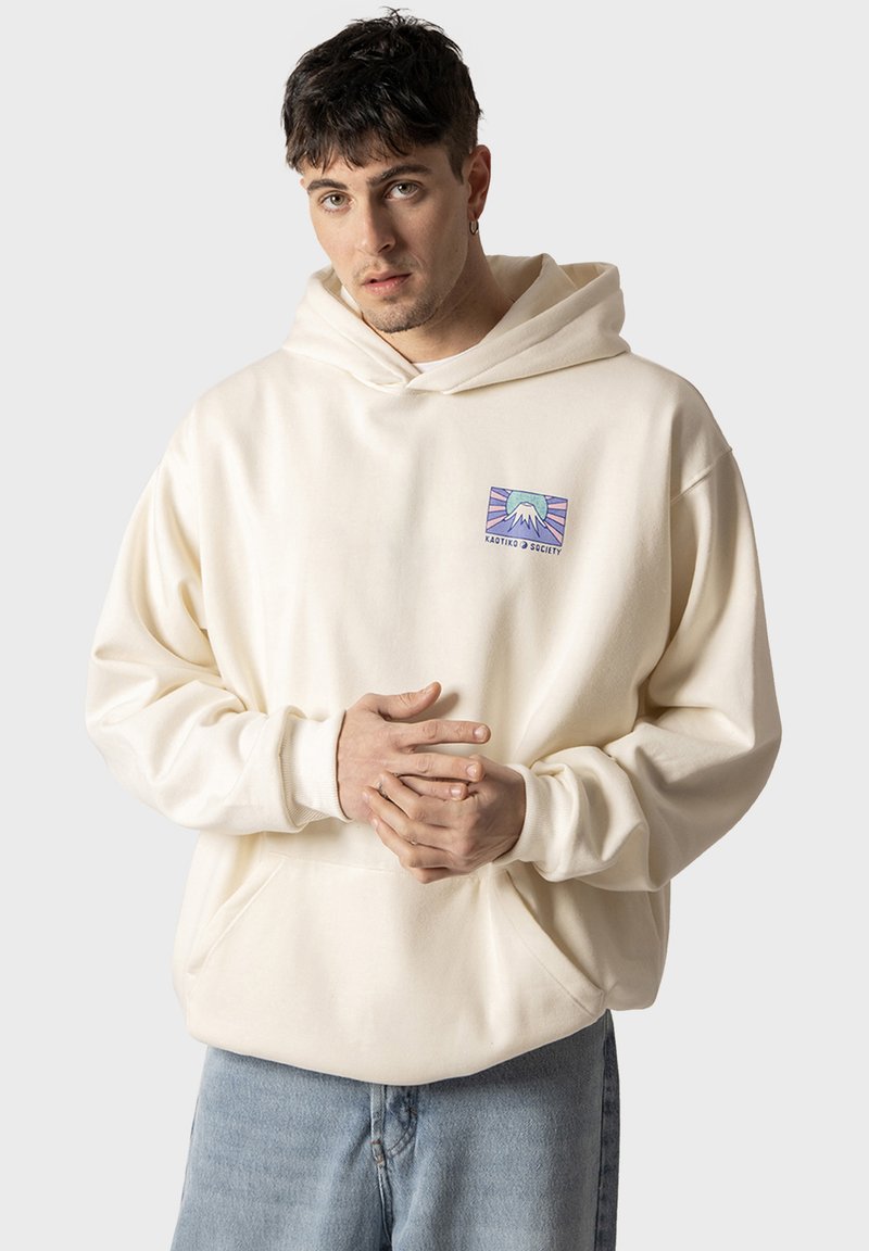 Sudadera con capucha de color crema, con un parche gráfico que presenta un diseño de montaña y texto, hecha de tela suave y con un corte relajado.