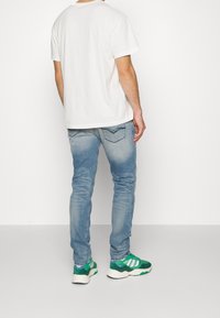 En man som bär en vit T-shirt, urtvättade blå jeans och gröna och vita sneakers, står mot en enkel vit bakgrund, med ryggen åt.
