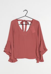 Blouse rose avec manches longues évasées et décolleté en V profond. Dos avec fermeture à nouer et deux lanières. Tissu lisse et léger.