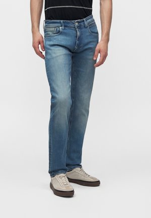 Férfi fakó kék slim-fit farmert és bézs sportcipőt visel, egy egyszerű fehér háttér előtt áll.