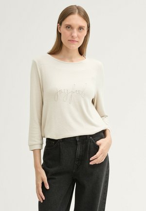 Top beige a maniche lunghe con il testo ricamato "joyful", caratterizzato da un colletto tondo e un tessuto morbido e testurizzato, abbinato a pantaloni neri a vita alta.