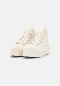 Białe wysokie sneakersy na platformie w odcieniu off-white, z grubymi teksturowanymi podeszwami, metalowymi oczkami i sznurowadłami na jednolitym białym tle.