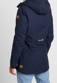 Mörkblå vadderad jacka med huva, dragsko i midjan och läderlogotypdetaljer. Har en slät yta och knäppta ärmslut.