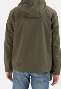 Veste imperméable vert olive avec capuche et détails sur les coutures latérales, associée à un jean bleu. Le tissu a une texture lisse et légère.