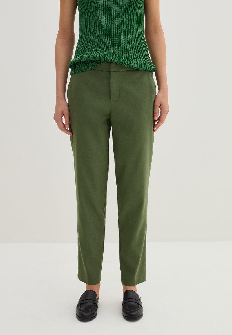 Grüne maßgeschneiderte Hose mit geradem Schnitt, einem glatten Material, seitlichen Taschen und einer mittelhohen Taille. Kombiniert mit schwarzen Slippern.