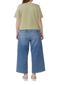 Olivgrünes Baumwoll-T-Shirt, kombiniert mit blauen, weit geschnittenen Jeans. Die Schuhe sind weiße Riemchensandalen. Einfaches, lässiges Design; keine sichtbaren Muster.