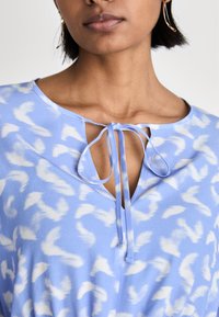 Bluză de culoare albastru deschis cu modele abstracte albe, guler rotund și detaliu de nod în față, fabricată dintr-un material moale cu o textură netedă.