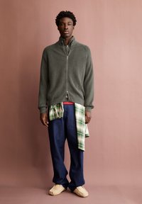 Dunkelgrüner Reißverschluss-Pullover mit gerippter Textur, kombiniert mit lockeren blauen Jeans. Grün-weißer Karoshirt um die Taille gebunden. Beigefarbene Schuhe.