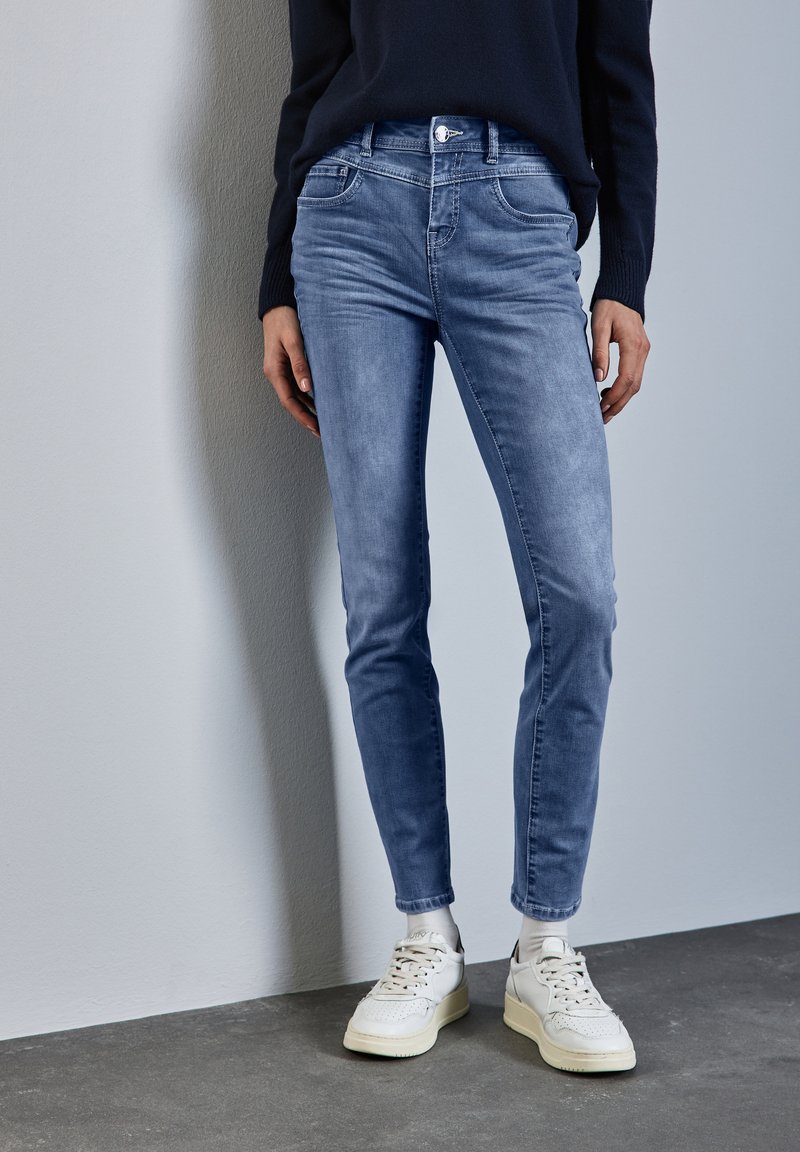 Ljusblå skinny jeans i denim, med en klassisk femfickedesign och metallknappstängning, kombinerade med vita sneakers.