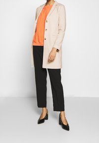 Cappotto lungo beige con bottoni dorati, abbinato a un maglione a costine arancione e pantaloni neri su misura. Tacchi neri a punta completano l'outfit.