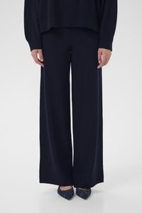 Άτομο που φοράει μαύρο παντελόνι με φαρδύ καβάλο και μαύρο μακρυμάνικο τοπ που ταιριάζουν, συνδυασμένο με σκούρα μυτερά παπούτσια με ορατό ράψιμο.