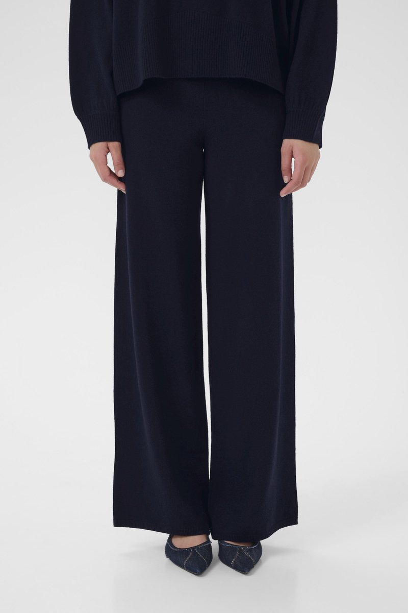 Άτομο που φοράει μαύρο παντελόνι με φαρδύ καβάλο και μαύρο μακρυμάνικο τοπ που ταιριάζουν, συνδυασμένο με σκούρα μυτερά παπούτσια με ορατό ράψιμο.