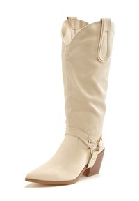 Bota de ante beige hasta la rodilla con punta afilada, tacón apilado y detalles decorativos de Hardware en el tobillo. Textura suave, diseño moderno.