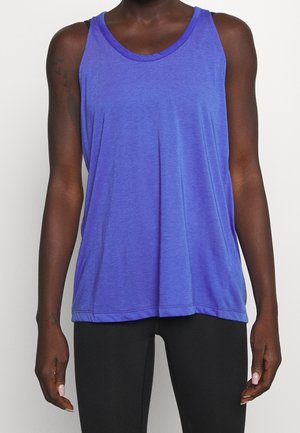 Personne portant un débardeur bleu sans manches et un pantalon noir ajusté, debout devant un fond clair uni.