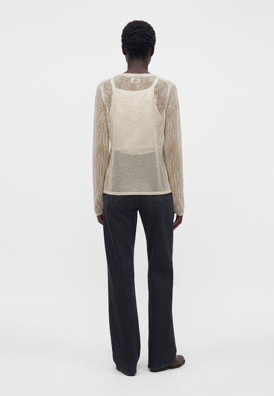 Personne aux cheveux courts et bouclés porte un haut à manches longues en maille beige par-dessus un débardeur, avec un pantalon noir à jambes larges, debout devant un fond uni.