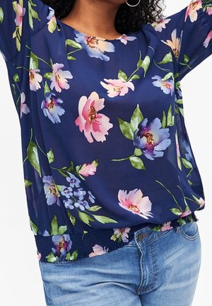 Femme portant un chemisier bleu marine à fleurs roses, violettes et blanches, associé à un jean en denim bleu clair, les bras levés au-dessus de la tête.