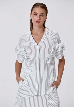 Jeune femme portant une chemise blanche à rayures verticales avec des volants sur les manches courtes, mains dans les poches, fond neutre.