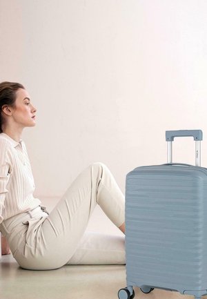 Mujer con atuendo beige sentada en el suelo reclinada hacia atrás, junto a una maleta rígida azul claro cerrada con asa extensible y ruedas.