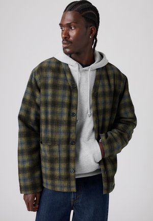 Homme aux cheveux tressés portant une veste à carreaux verte par-dessus un sweat à capuche gris et un jean bleu, regardant vers sa gauche avec une main dans la poche.