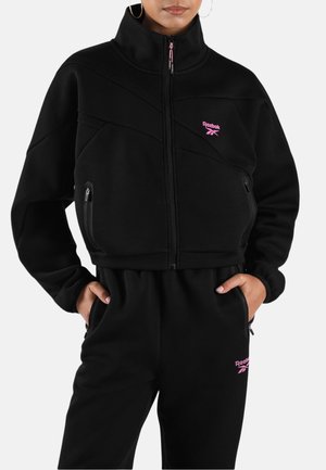 Frau trägt schwarzen Reebok Trainingsanzug mit pinkem Logo, Jacke mit Reißverschluss und passenden Hosen, Hände in den Taschen, steht vor weißem Hintergrund.