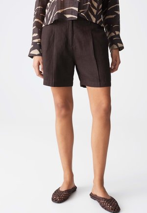 Femme portant un short taille haute sur mesure marron foncé, un haut à manches longues à motif zèbre et des chaussures tissées marron sans lacets, debout devant un fond blanc.