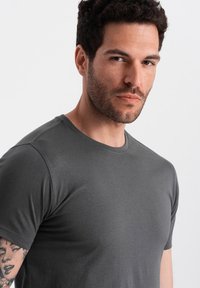 Szara koszulka z krótkim rękawem wykonana z mieszanki bawełny, z okrągłym dekoltem i gładką fakturą. Model ma widoczny tatuż na ramieniu.