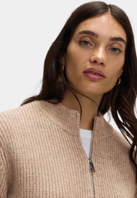 Béžová pletená bunda s vysokým límcem a stříbrným zipem. Má strukturovanou tkaninu a je nošena přes bílou blůzu.