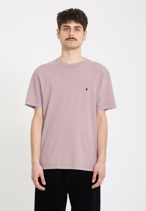 Jeune homme aux cheveux bruns courts et moustache, portant un t-shirt violet clair et un pantalon noir, debout devant un fond blanc uni.