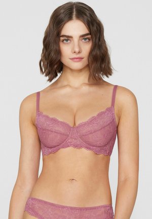 Femme aux cheveux bruns mi-longs portant un soutien-gorge et une culotte en dentelle rose assortis, face à un fond uni.