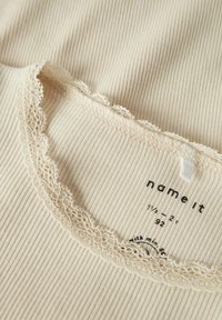 Kremowy materiał w prążki z koronkowym wykończeniem wzdłuż dekoltu, posiadający metkę z informacjami o rozmiarze i nazwą marki "name it."